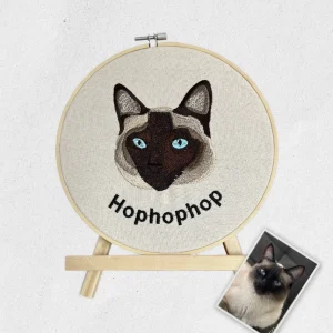 Portrait de chat Siamois brodé personnalisé sur un tambour en bois sur son chevalet avec la photo d'origine posée à côté.