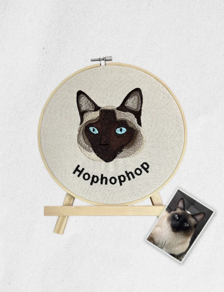 Portrait de chat Siamois brodé personnalisé sur un tambour en bois sur son chevalet avec la photo d'origine posée à côté.