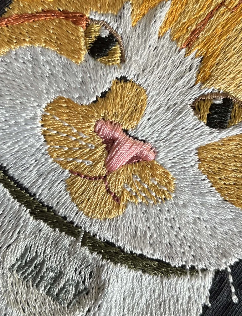 Gros plan macro sur une broderie détaillée de visage de chat montrant la texture des fils blancs, roux et roses, les points de contour et le relief des moustaches sur tissu noir.