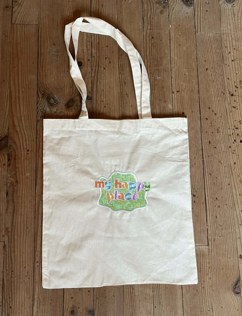 Sac tote-bag en coton naturel beige posé sur un parquet en bois, personnalisé avec un écusson brodé carré vert affichant le texte coloré "my happy place".