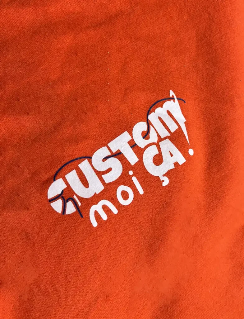 Gros plan macro sur un textile orange vif avec le logo de la marque "Custom moi ça!" imprimé en blanc et noir, posé sur une surface texturée.