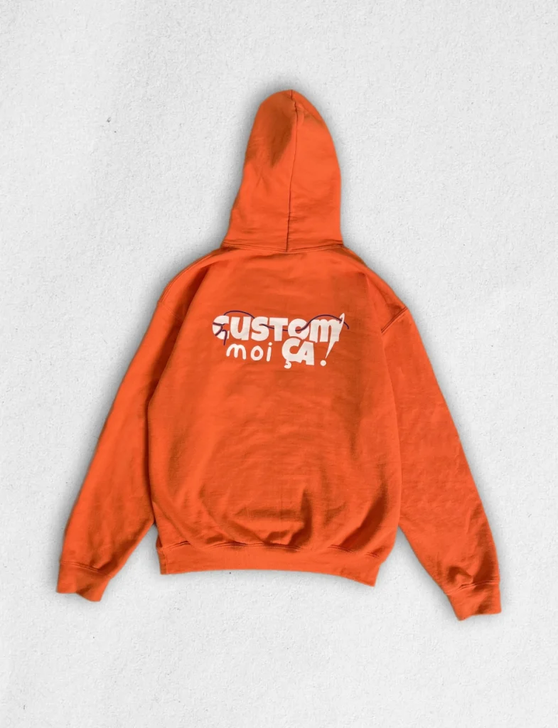 Sweat à capuche orange vif personnalisé avec le logo "Custom moi ça !" imprimé en blanc et noir sur le dos, posé à plat sur fond clair.