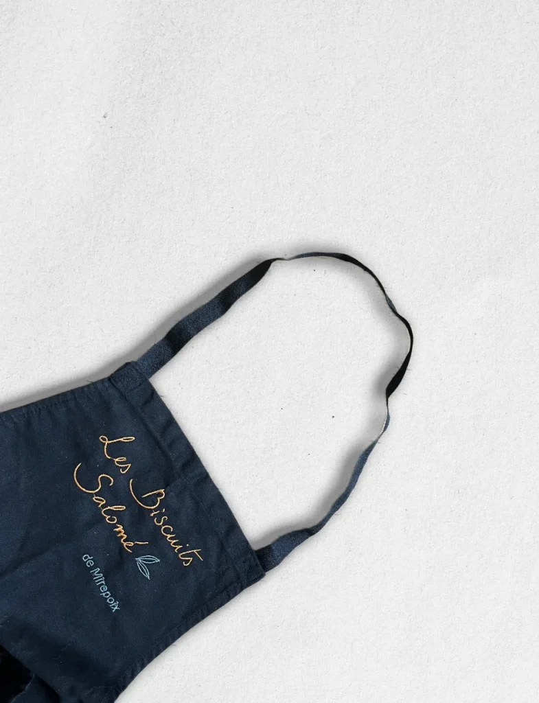 Tablier professionnel noir avec logo "Les Biscuits Salomé de Mirepoix" brodé en fil doré, vue rapprochée sur la sangle et le haut du tablier.