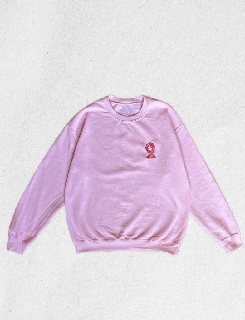 Sweat rose pastel sans col posé à plat, avec un ruban Octobre Rose brodé de fleurs de sakura sur le cœur